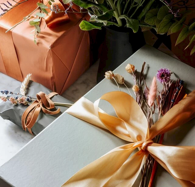 The image shows beautifully wrapped gift boxes adorned with ribbon and dried flowers, set on a table beside a vase with green foliage.