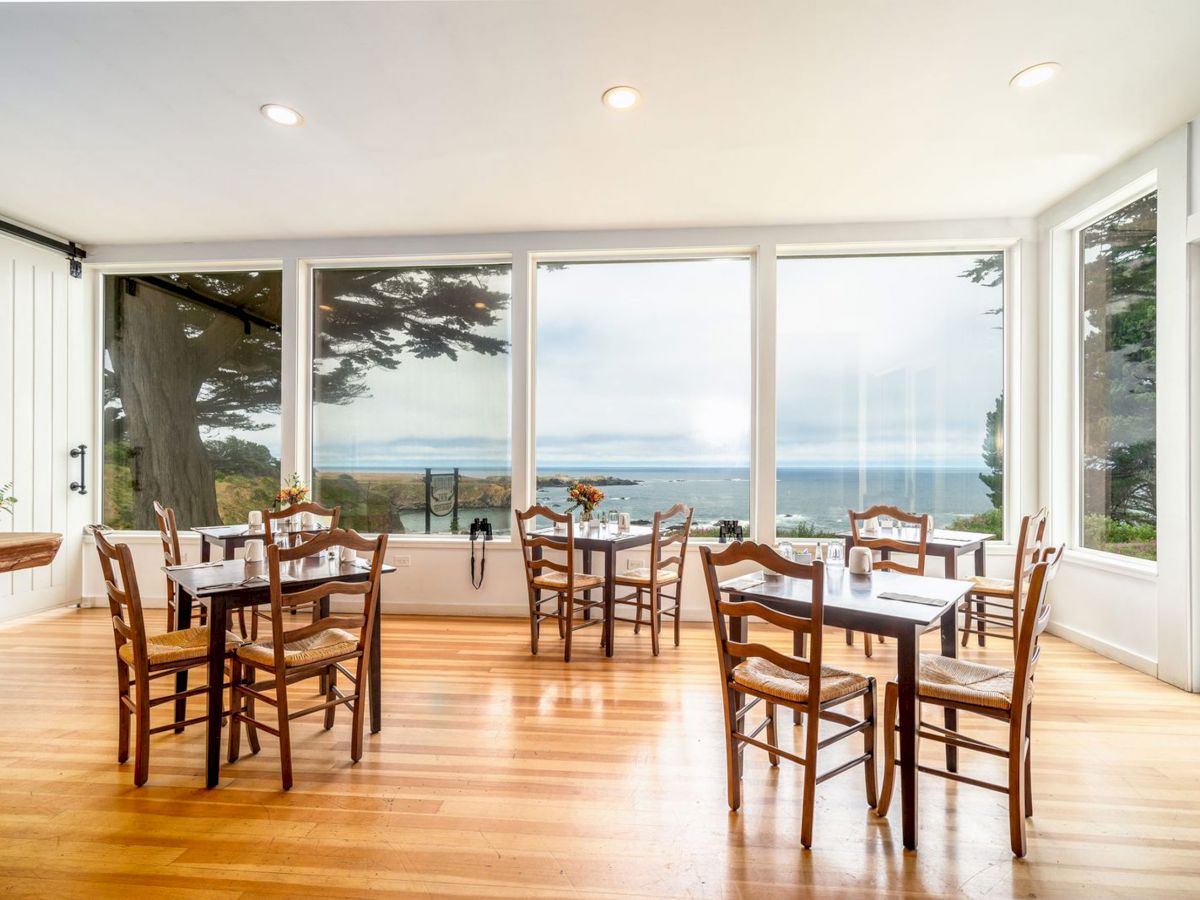 The image shows a bright, airy dining room with wooden chairs and tables, large windows offering a beautiful ocean view, and a light wood floor.