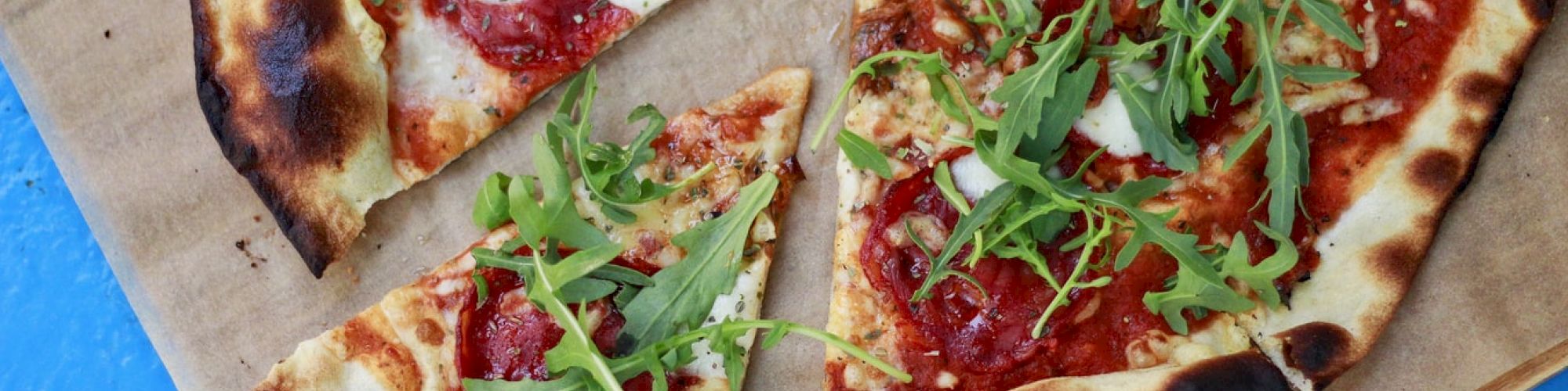 A pizza with tomato sauce, cheese, and fresh arugula on top, placed on brown paper on a blue surface, with one slice partially removed.
