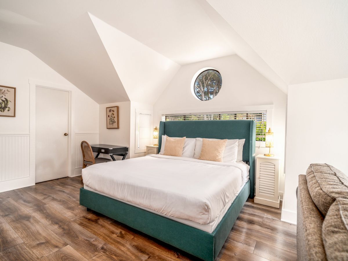 A cozy bedroom with a teal bed, white bedding, wooden floors, a desk, and wall artwork under a sloped ceiling and round window ending the sentence.