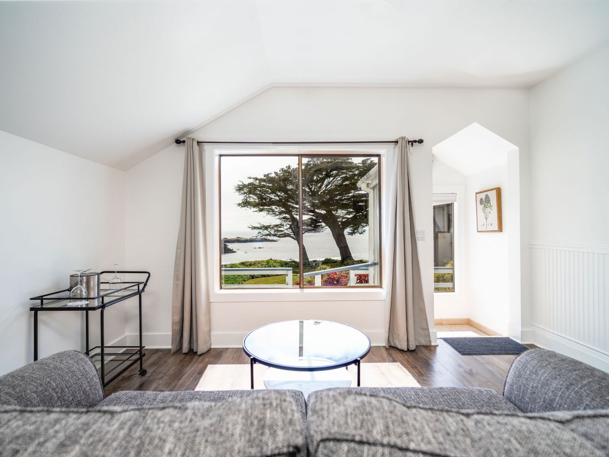 The image shows a bright living room with a large window, grey sofa, glass table, and a scenic ocean and tree view outside.