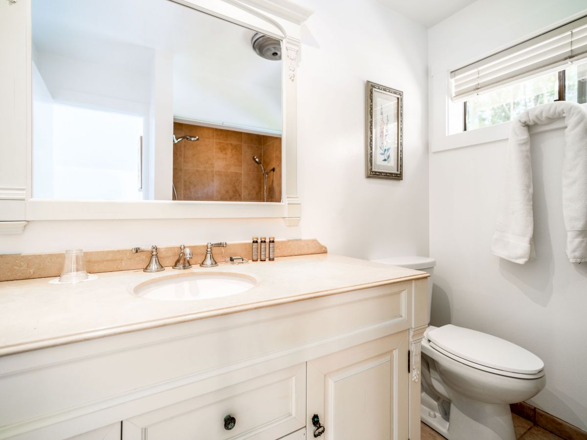 A bright bathroom features a large mirror, sink, countertop, toilet, and towels hanging beside a window with blinds, ending the sentence.