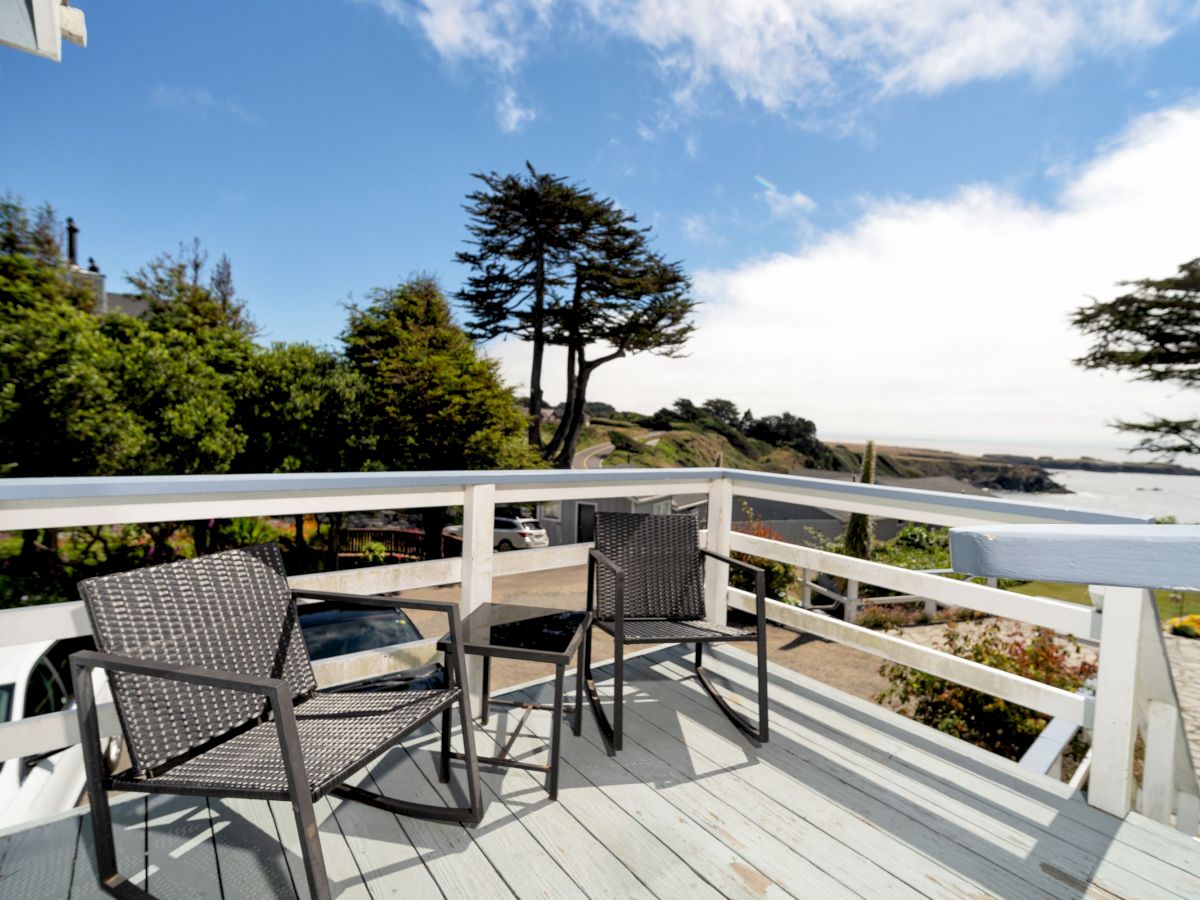 A wooden deck with two chairs offers a scenic view of a coastline, with trees and blue skies in the background, overlooking the ocean.