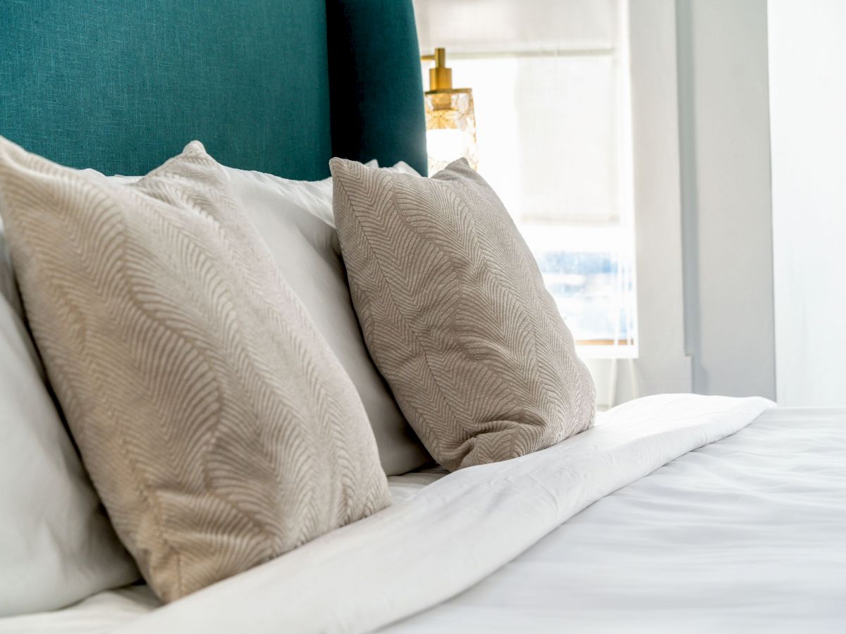 The image shows a neatly made bed with two beige, patterned pillows against a teal headboard in a bright, well-lit room.