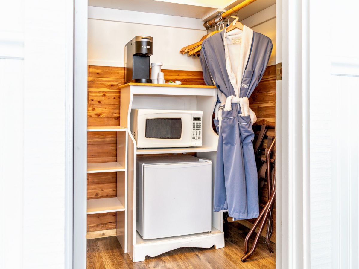 A compact closet setup with a coffee maker, microwave, mini refrigerator, hanging robe, and an iron on an ironing board, neatly organized.