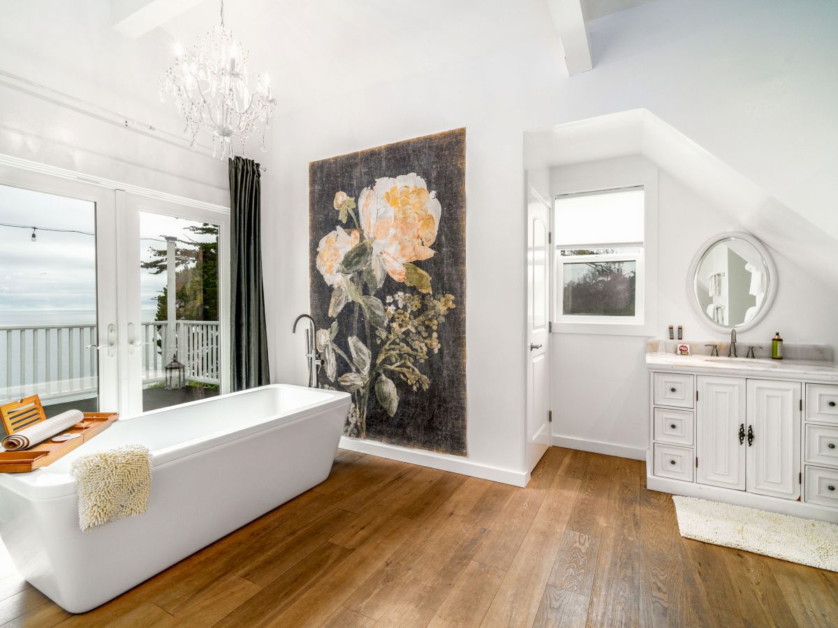 A modern bathroom with a freestanding tub, chandelier, large floral artwork, vanity with round mirror, wood flooring, and natural light from windows.