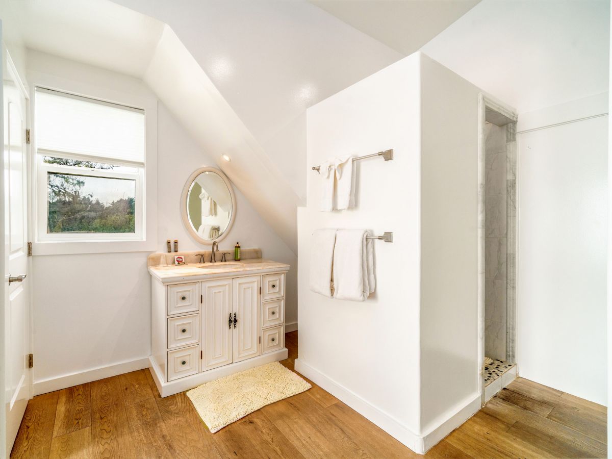 A bright bathroom with a vanity, round mirror, towels on racks, a small rug, and a standalone shower on hardwood floors.