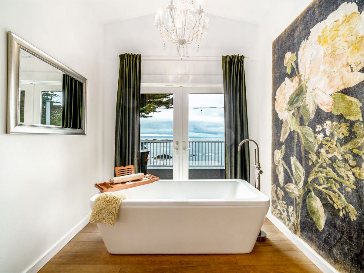 A modern bathroom features a freestanding bathtub, a large floral mural, a chandelier, and a view of the balcony through glass doors.