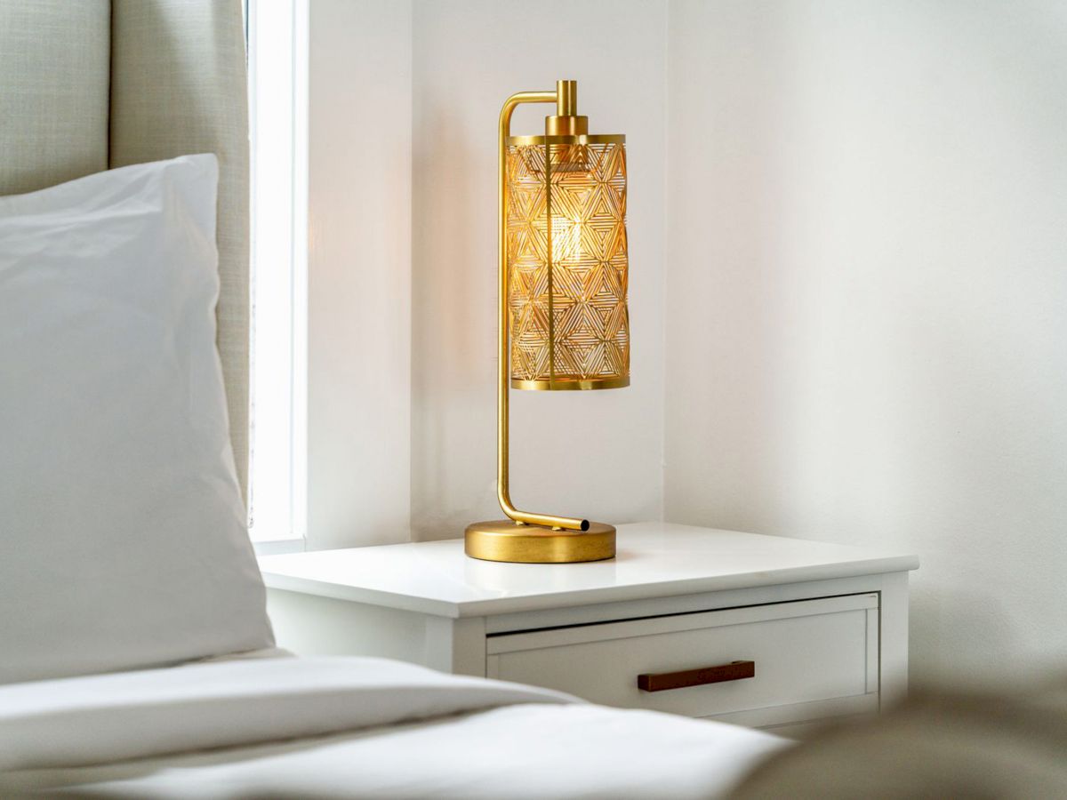 A lit decorative lamp sits on a white bedside table next to a bed with white linens in a minimalist bedroom.