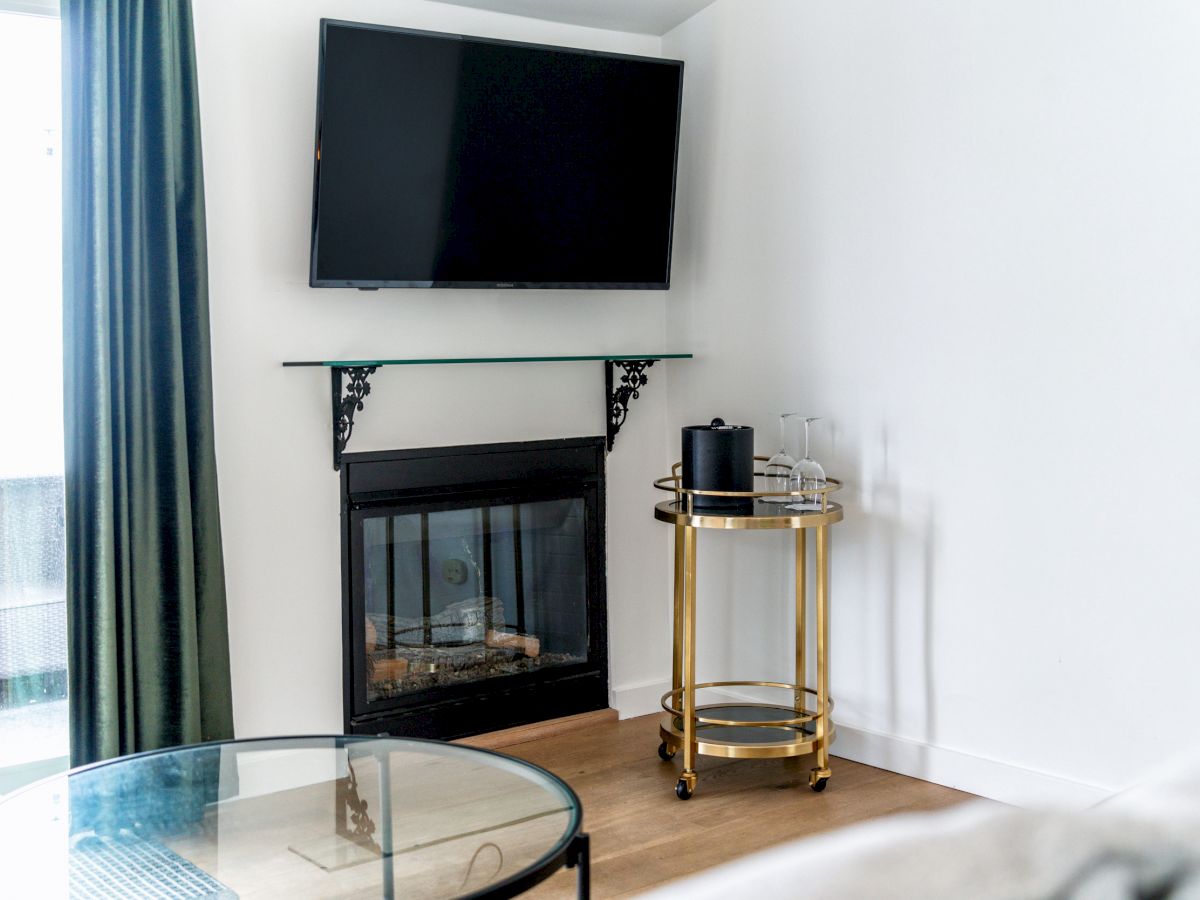 A modern living area with a wall-mounted TV above a fireplace, a round glass coffee table, and a gold bar cart with a speaker and glasses.