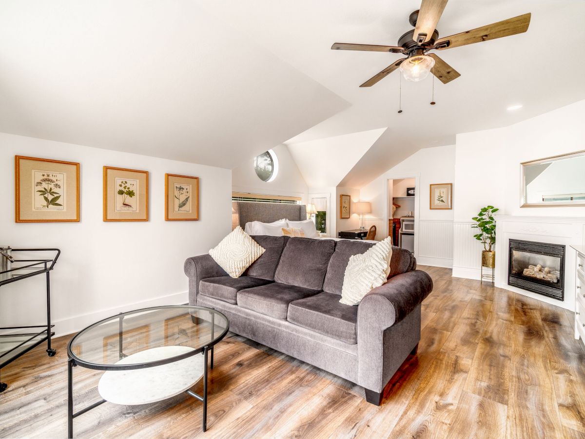 A cozy living room features a grey couch, a round glass coffee table, framed artwork, a ceiling fan, and a fireplace, with an open kitchen in the back.