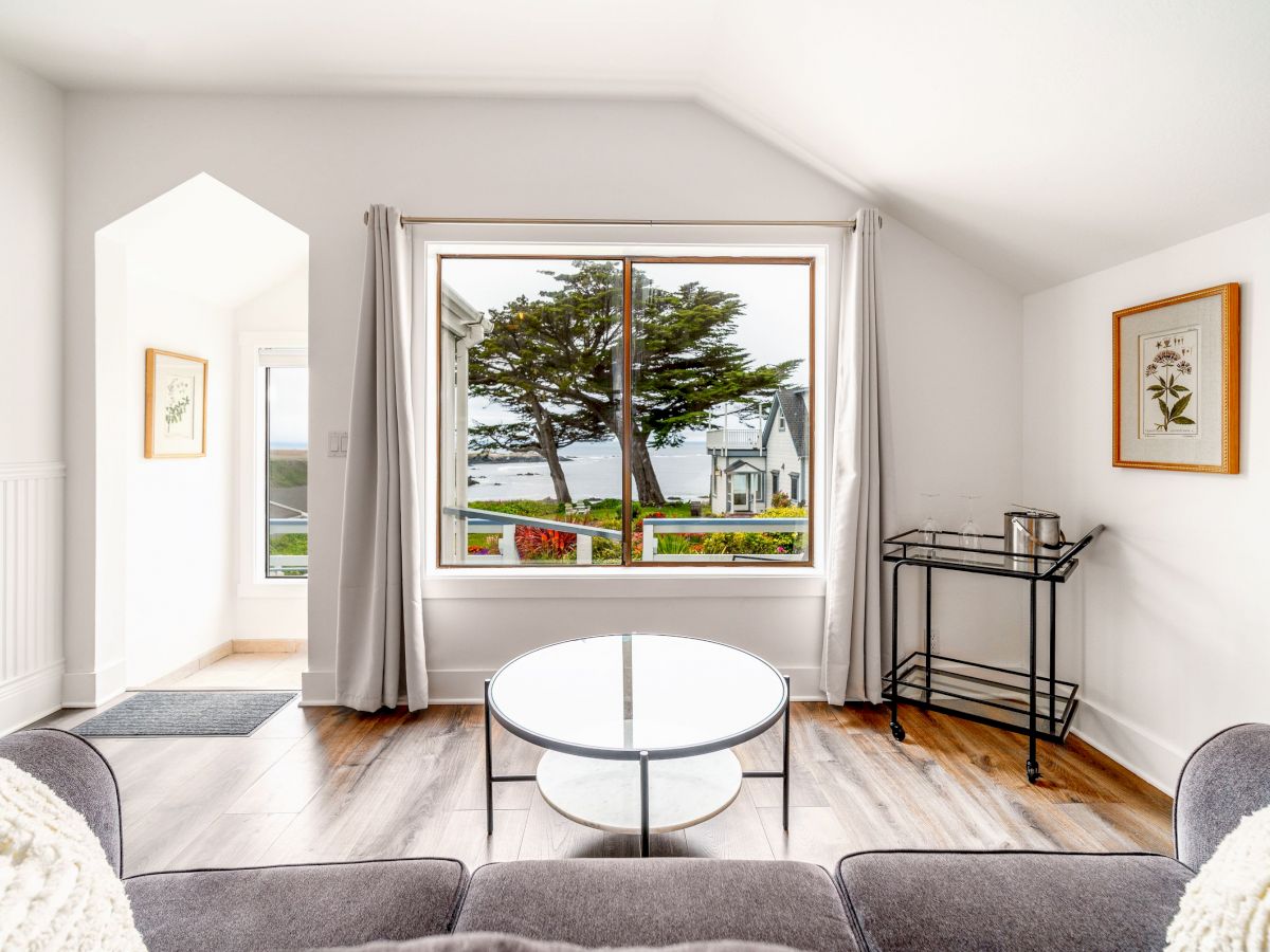 A cozy living room with a large window offering a scenic view of trees and water, featuring a round coffee table, sofa, and wall art.