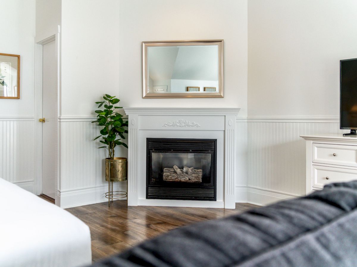 This image shows a cozy, modern living room with a fireplace, a mirror above it, a potted plant, a TV, and white furniture and walls.