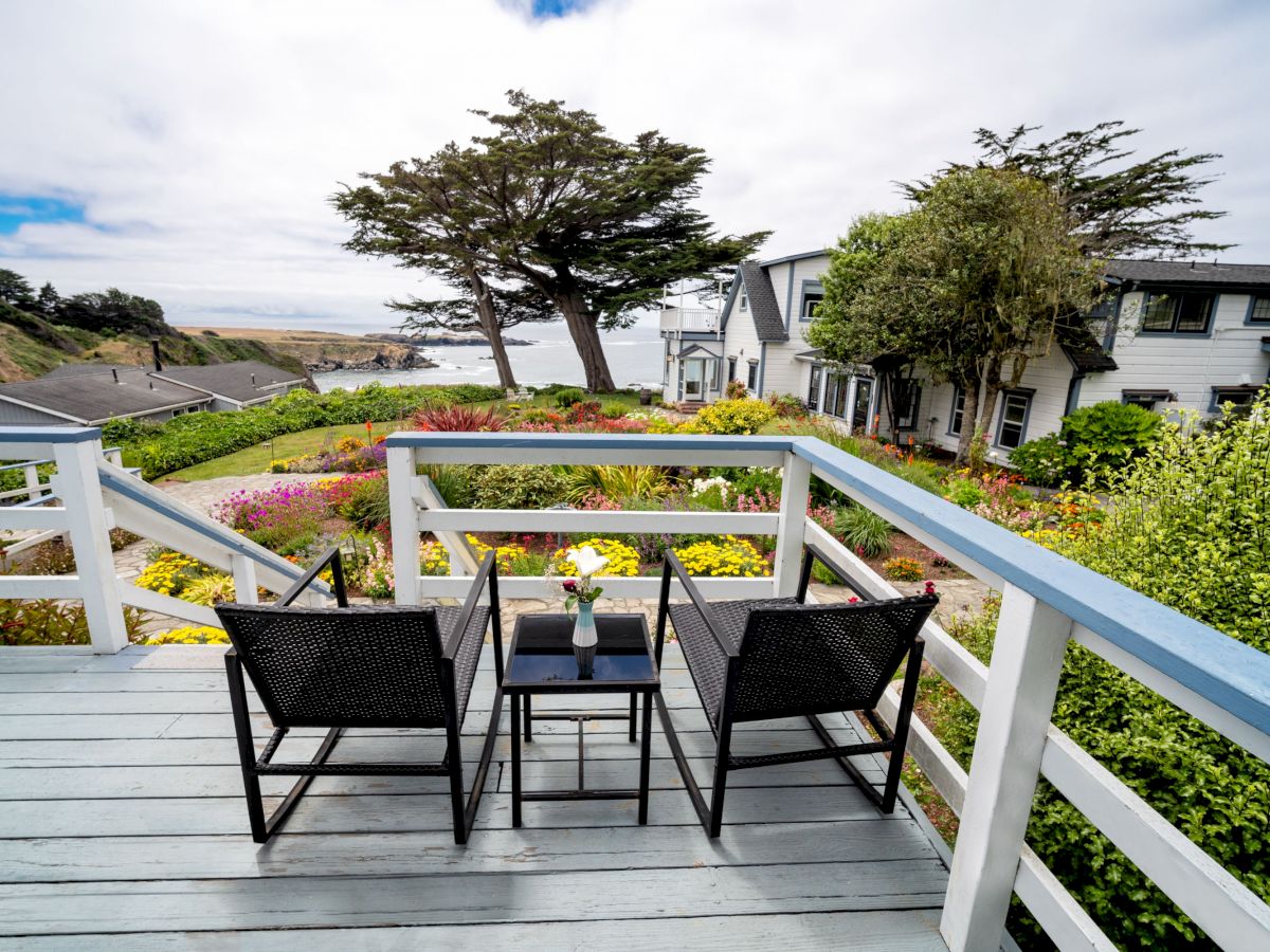 A cozy outdoor deck with two chairs and a table overlooking a colorful garden, houses, and a scenic ocean view in the background, under a partly cloudy sky.