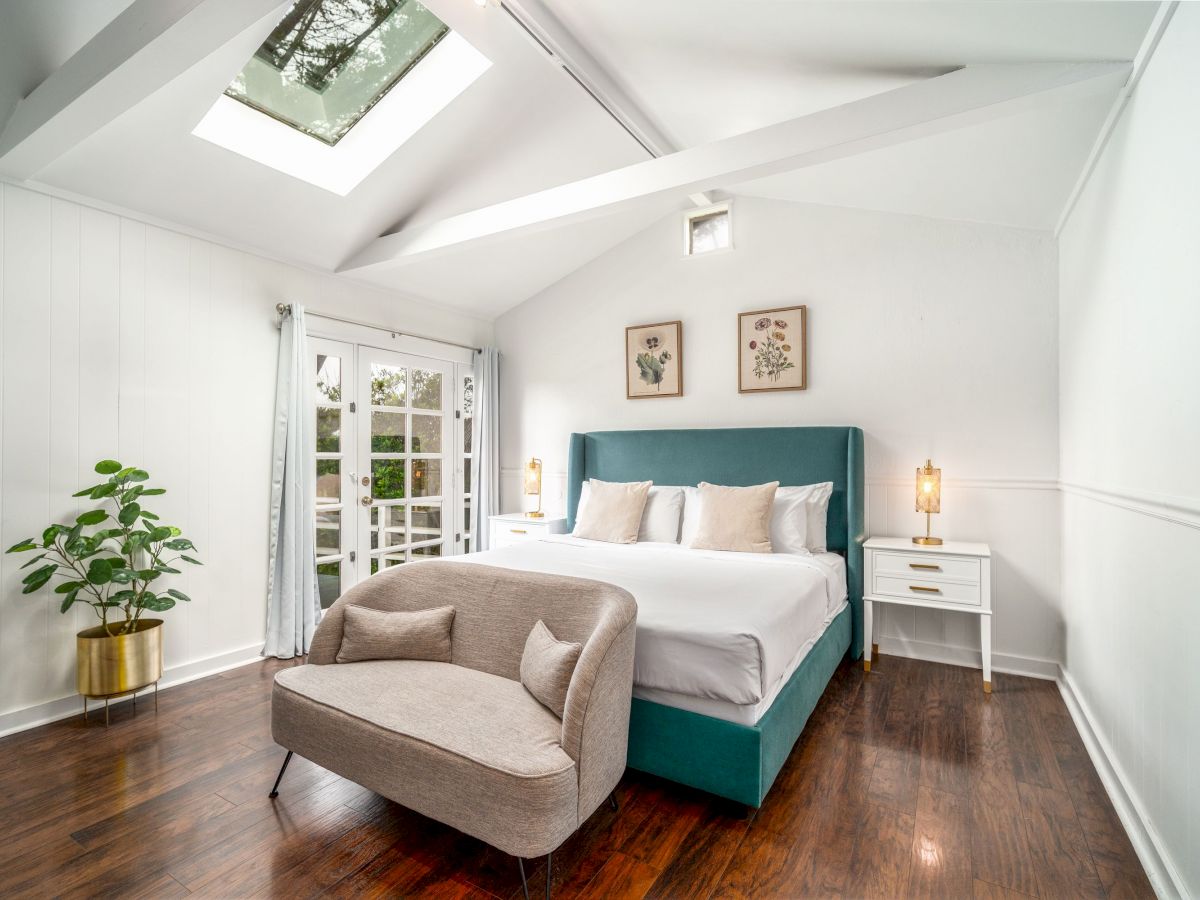 A bright bedroom featuring a skylight, a teal bed, a gray couch, white side tables, plants, and decor. French doors lead outside.