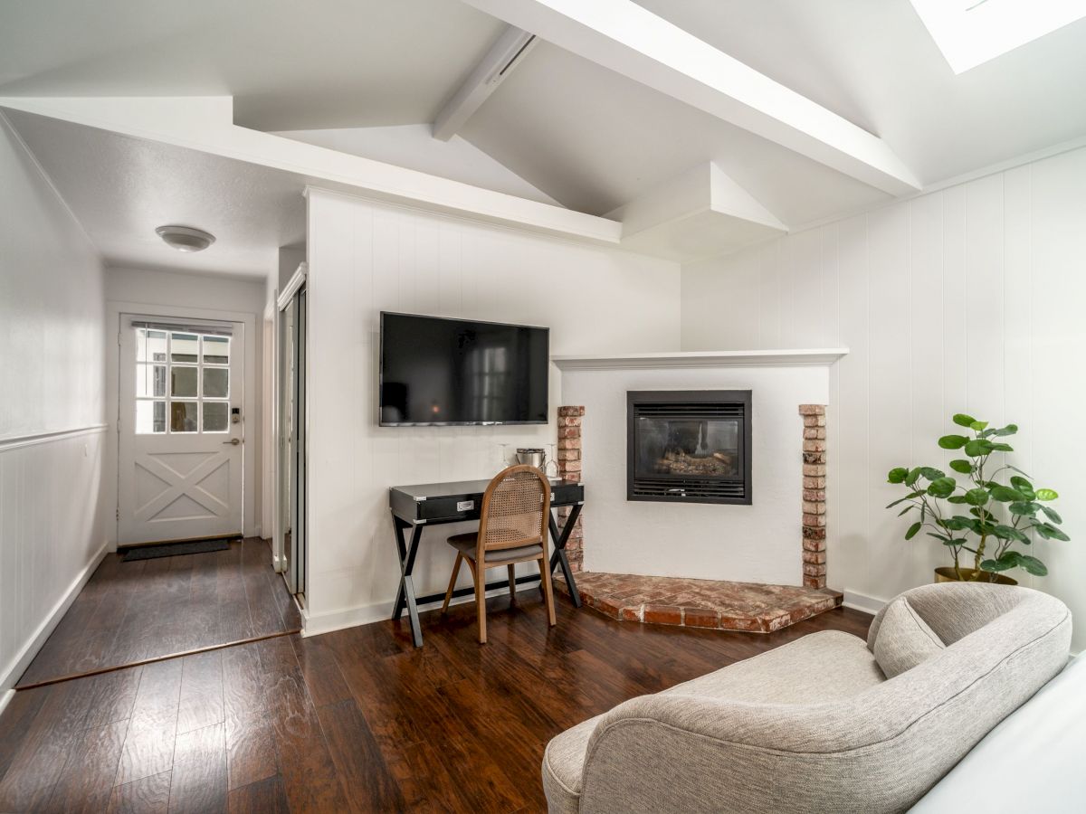 A cozy living area with white walls features a mounted TV, a small desk, a fireplace, a plant, and a couch, with wooden flooring throughout.