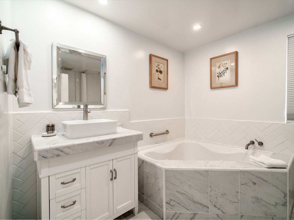 A bathroom with a white vanity, rectangular mirror, towel rack, bathtub with marble tiles, two framed artworks, and a window with blinds.