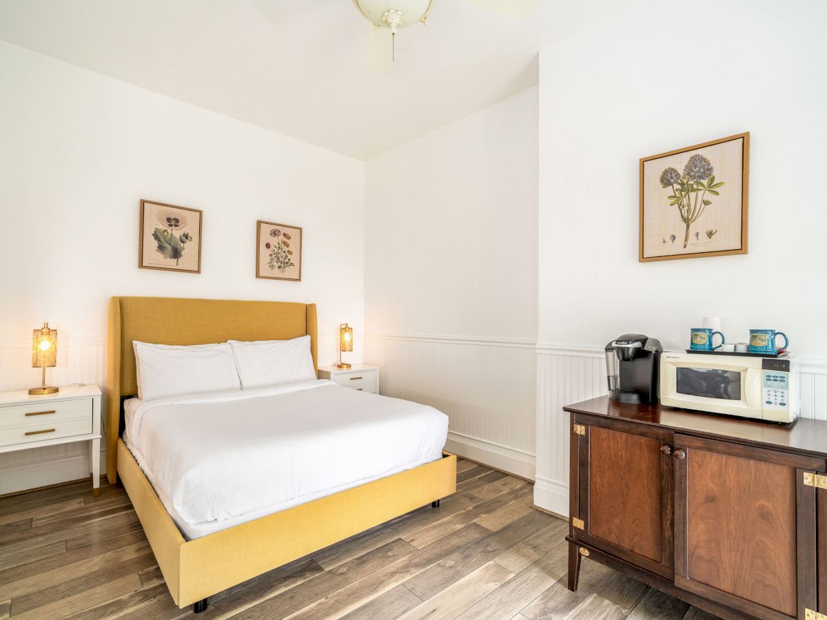 A cozy bedroom featuring a yellow bed, two side tables with lamps, a cabinet with a coffee maker and microwave, and framed wall art.