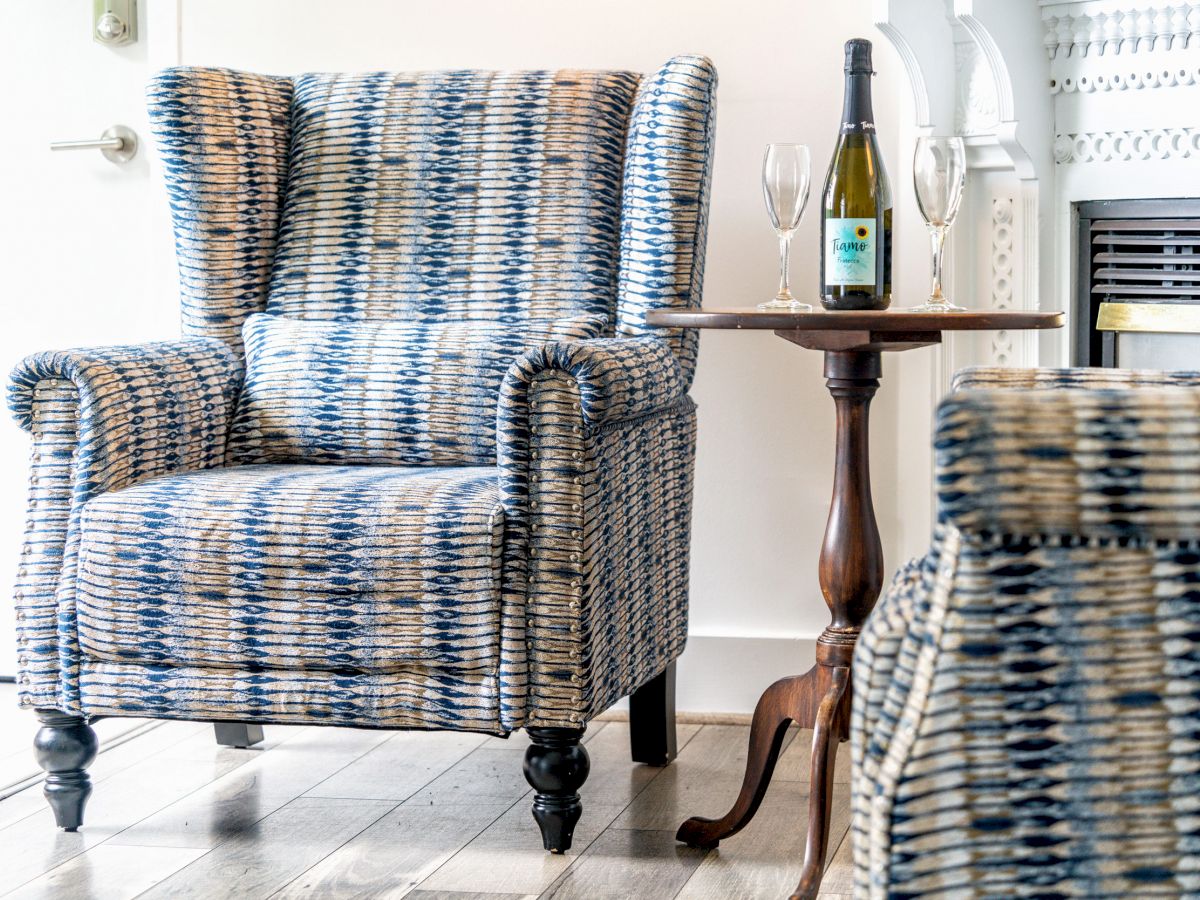 A cozy reading nook with a patterned armchair, a small round table with a bottle of wine and two glasses, and part of a fireplace visible in the background.