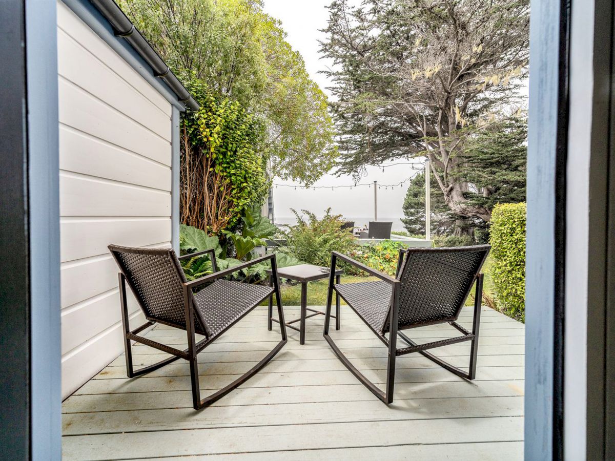 Two black rocking chairs and a small table on a wooden deck overlook a garden with trees and shrubs, seen through an open doorway.