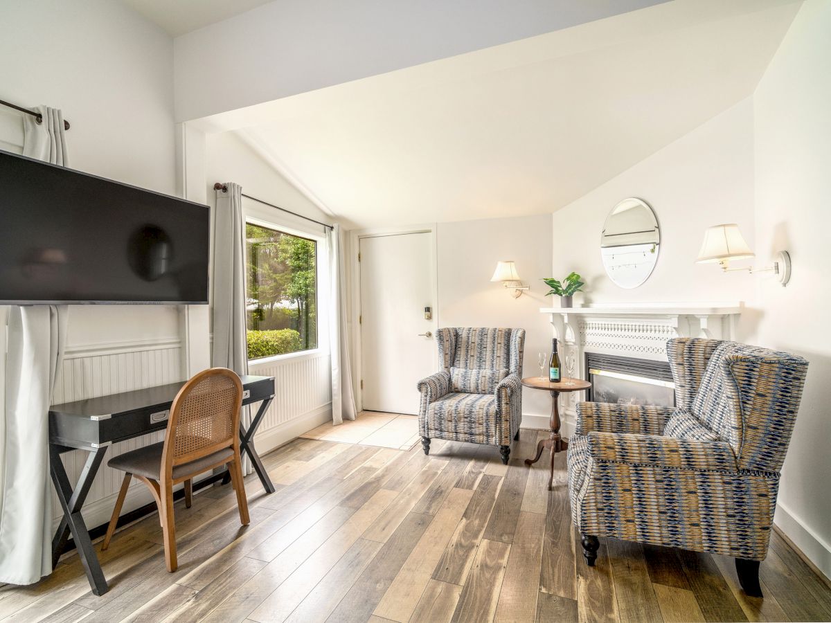 A cozy room with two patterned chairs, a small table, a wall-mounted TV, a writing desk, a large window, and a door.