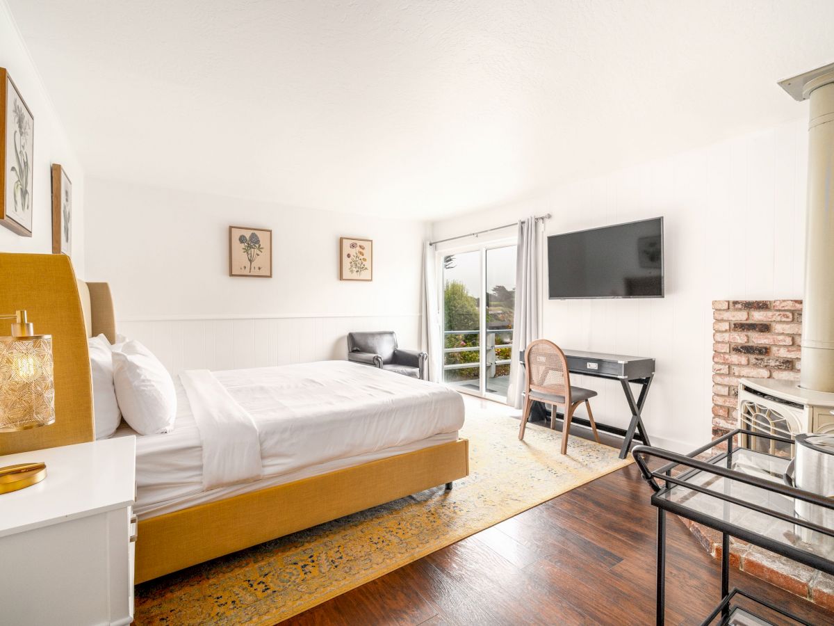 A bright bedroom with a large bed, wall art, a TV, a desk with a chair, a brick fireplace, and a window leading to a small balcony.