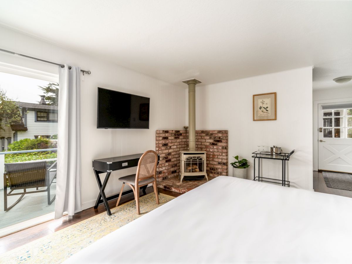 A cozy bedroom with a white interior, a wall-mounted TV, a desk, and a chair in front of a fireplace, adjacent to a sliding door leading outside.