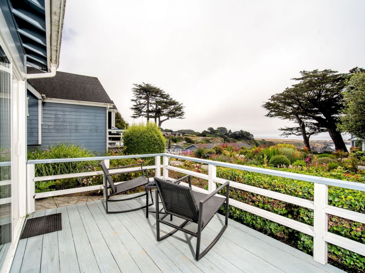 A cozy outdoor patio with two chairs and a table overlooks a lush garden and distant trees, with houses and a misty landscape in the background.