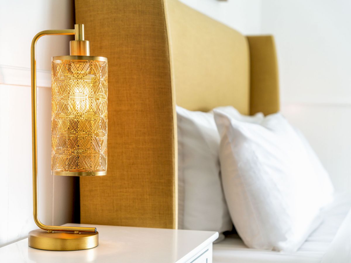 A stylish bedside area with a decorative lamp on a white nightstand, next to a bed with a golden headboard and white pillows.