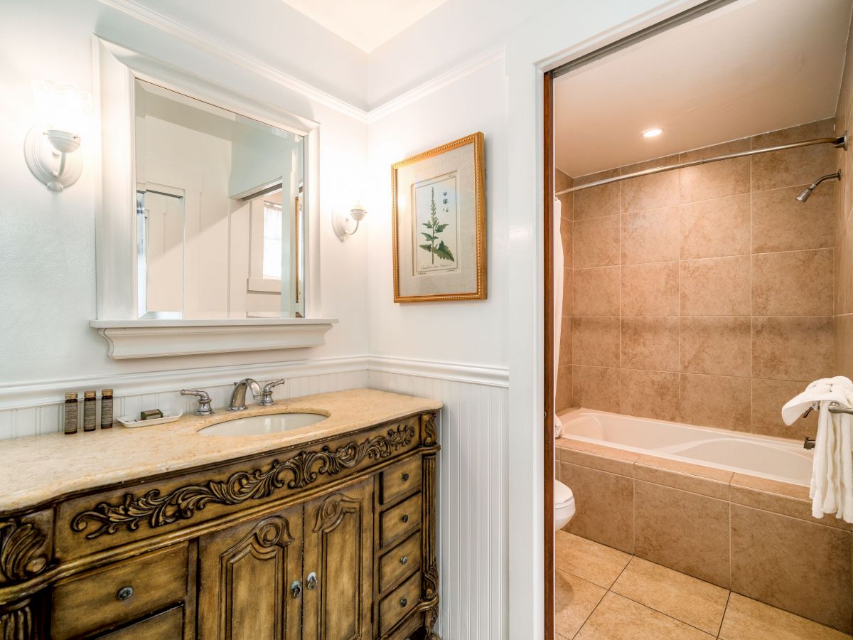 A bathroom with an ornate wooden vanity, a large mirror above the sink, a framed picture on the wall, and a separate tiled bathtub and shower area.