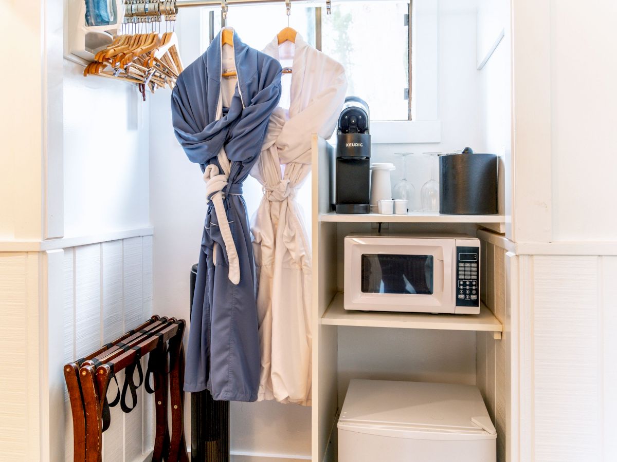 Two bathrobes, a coffee machine, a microwave, and a mini-fridge are in a small closet space with hangers and luggage racks placed nearby.