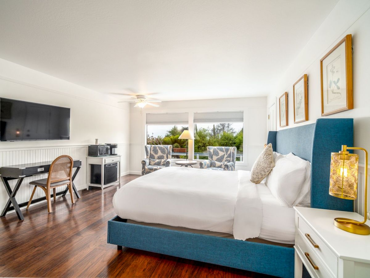 A modern bedroom with a large bed, wall-mounted TV, desk, and seating area by the window, overlooking an outdoor view.