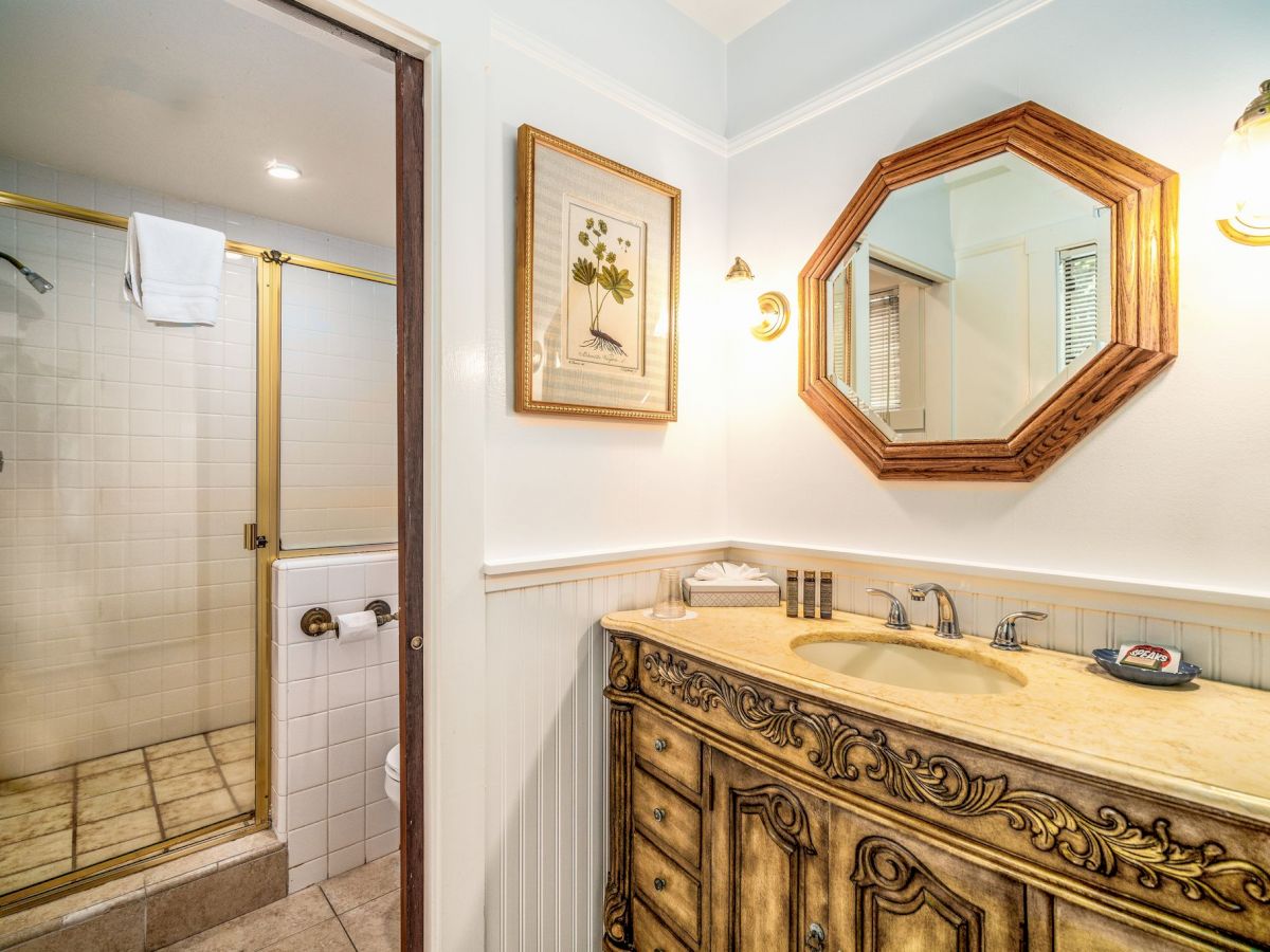 A bathroom features a shower with a glass door, a decorative vanity with a sink, a wall mirror, and framed art above white wainscoting, ending the sentence.