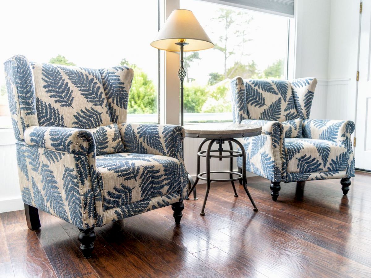 Two patterned armchairs and a small round table with a lamp between them sit near a window on wooden flooring.