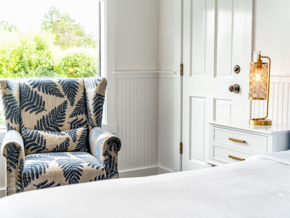 A cozy bedroom features a fern-patterned armchair, a white nightstand with a golden lamp, a bed with white linens, and a door.