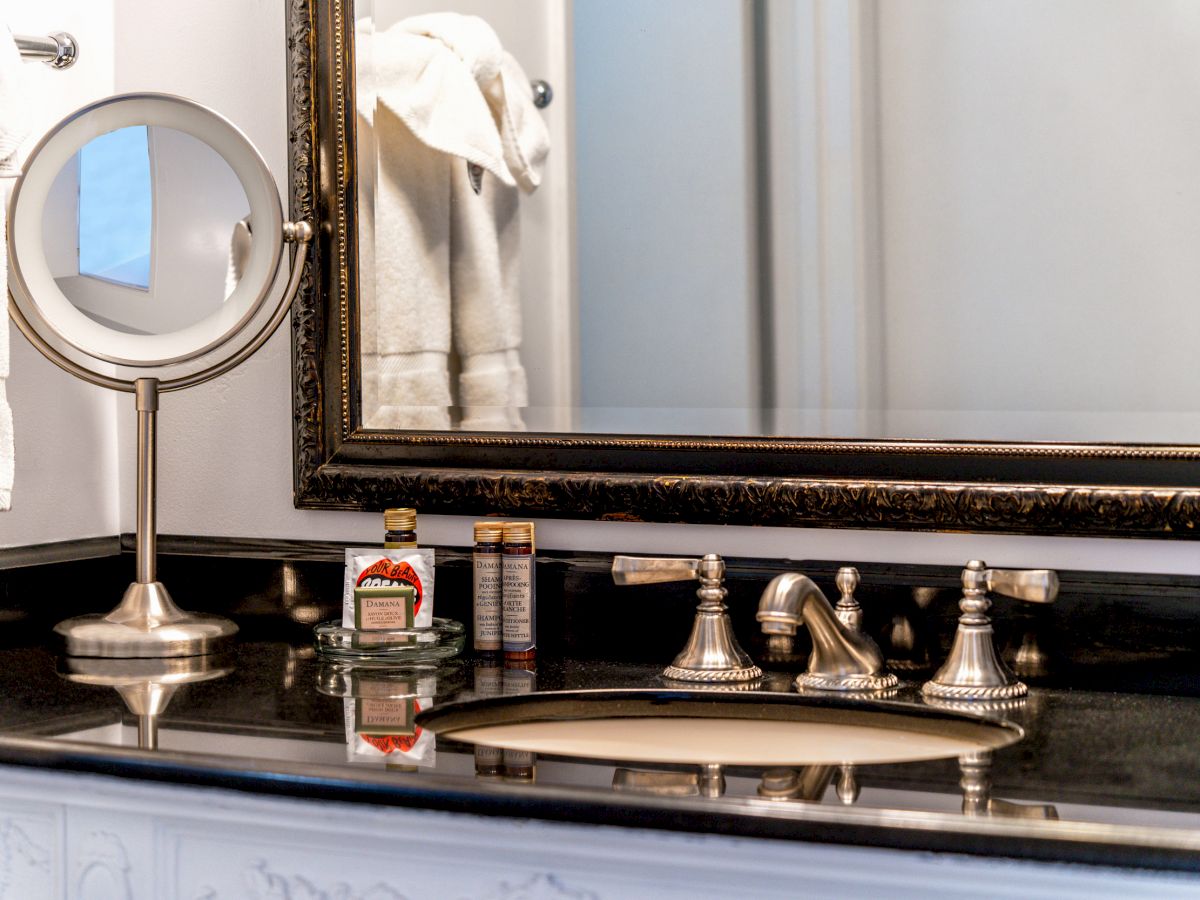 A bathroom countertop with a vanity mirror, a bottle of mouthwash, a grooming product, a sink with ornate fixtures, and towels hanging on the wall.