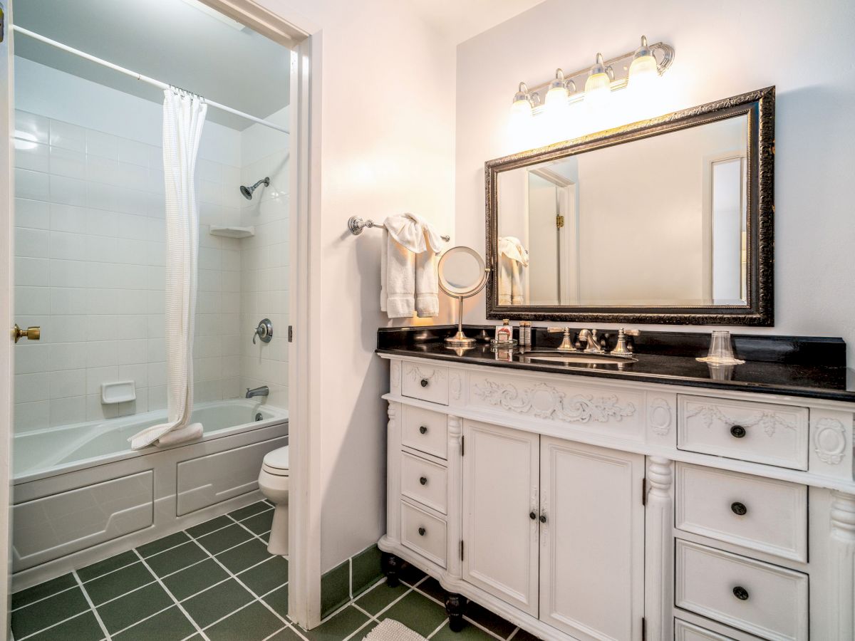 This image shows a clean bathroom with a tub-shower combo, white vanity, large mirror, tiled floor, and wall-mounted light above the sink.