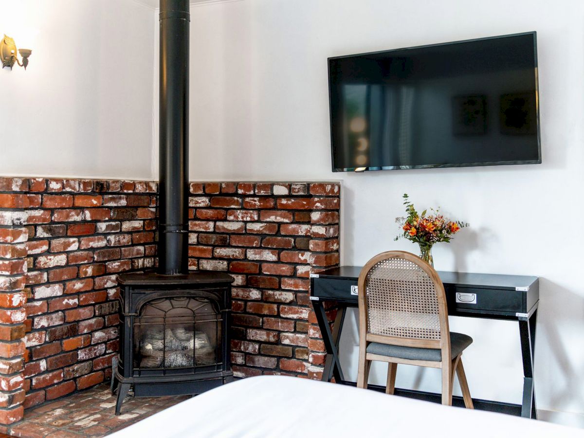 The image shows a cozy room with a brick fireplace, a black stove, a wall-mounted TV, a desk with a wicker chair, and a vase of flowers on the desk.