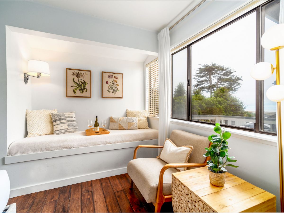 A cozy reading nook with a built-in bench, pillows, a chair, and a side table. Decor includes framed art, wall lamp, and potted plant by the window.