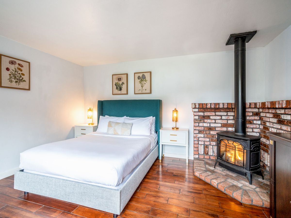 A cozy bedroom with a bed, framed art on the walls, nightstands with lamps, wooden floor, and a corner brick fireplace with a lit wood stove.