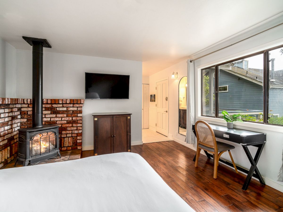 A cozy bedroom with a fireplace, wall-mounted TV, desk by the window, armoire, and wooden floors. The room looks bright and inviting.