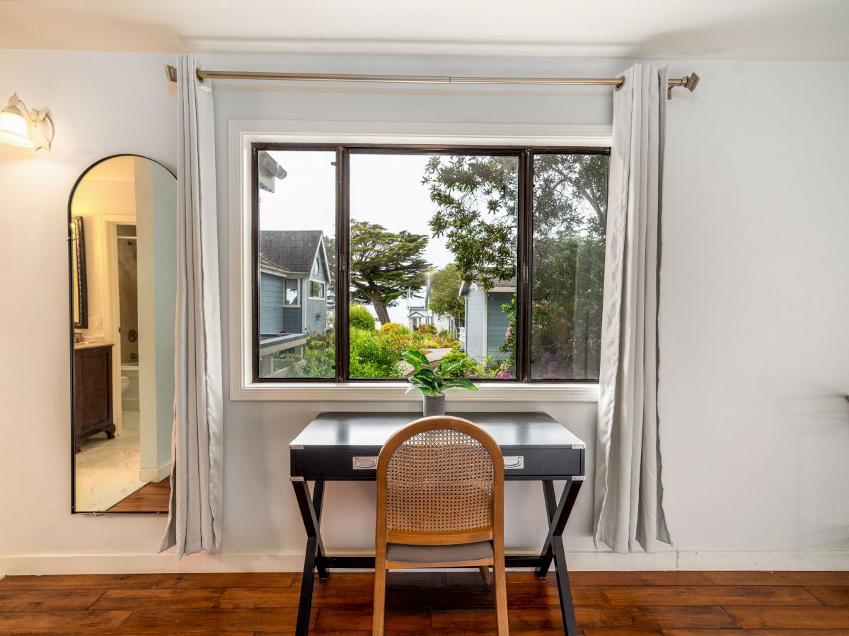 A modern desk with a chair facing a large window with a scenic view, next to a tall mirror on the left wall and white curtains, is seen in the image.