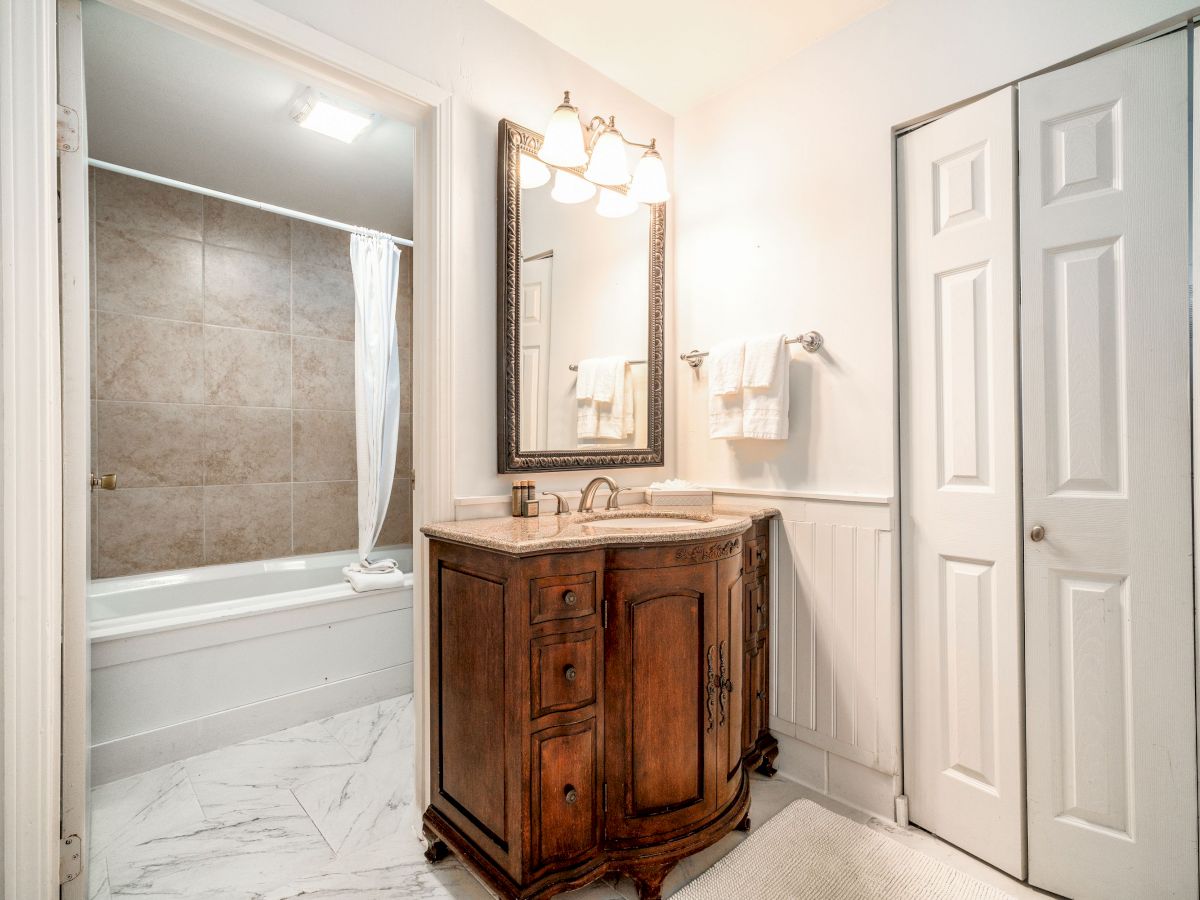 The image shows a bathroom with a wooden vanity, a large mirror, a bathtub with a curtain, white tiles, and a closet with sliding doors.