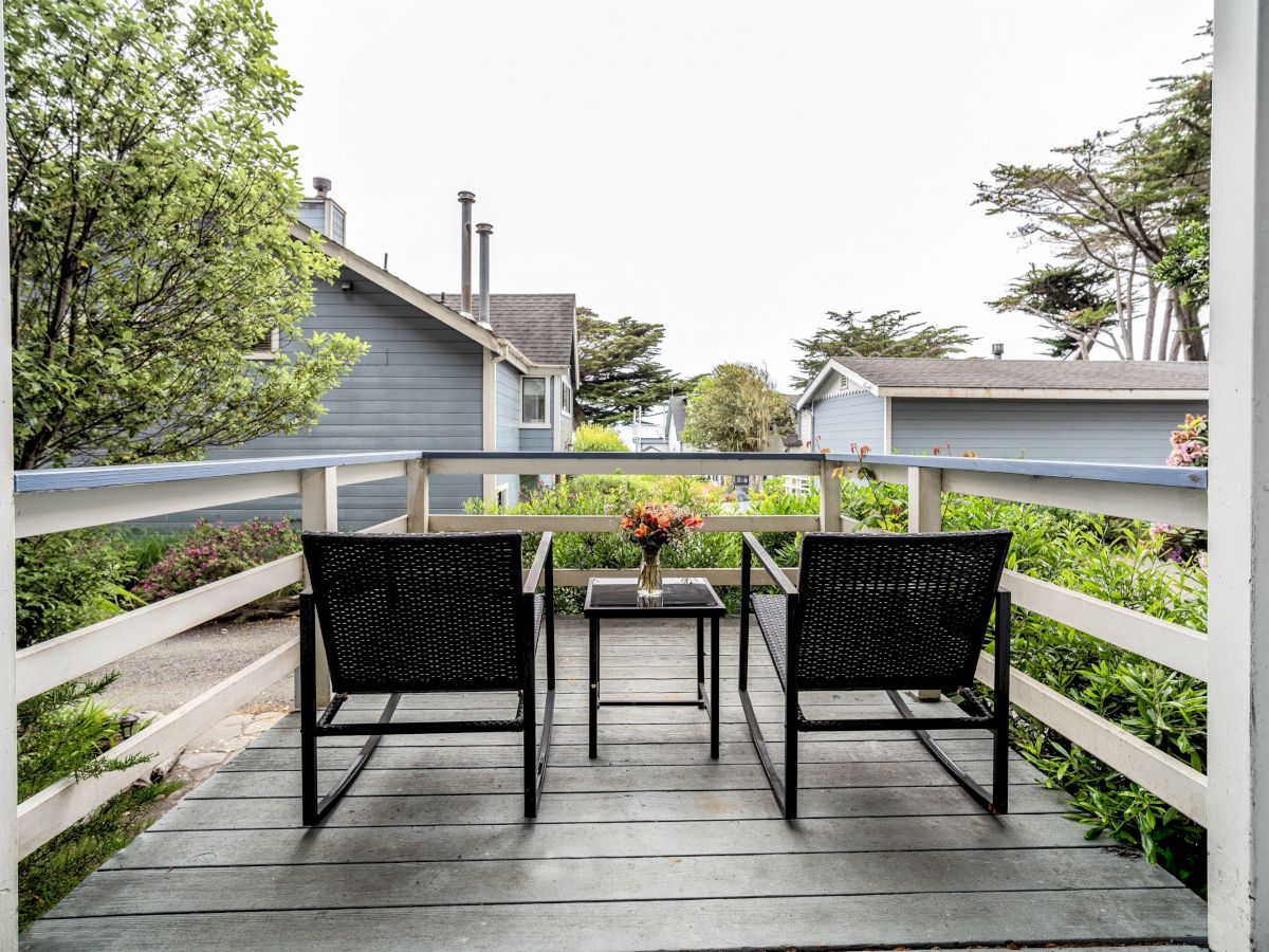 A small deck with two black wicker chairs and a table, featuring a flower vase, overlooks neighboring houses and trees in the background.