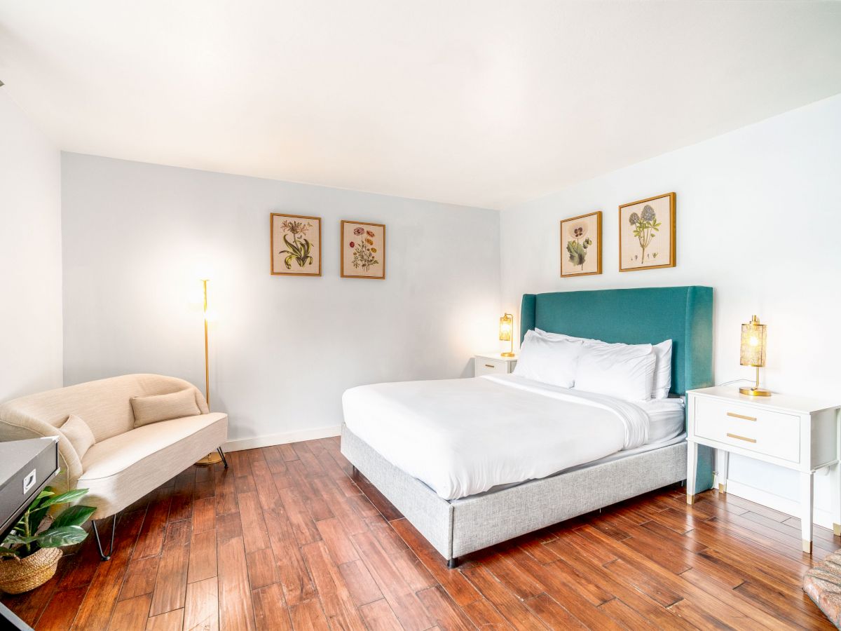 A bright bedroom with a bed, green headboard, nightstands, a beige loveseat, wooden floor, and framed art on the wall, ending the sentence.