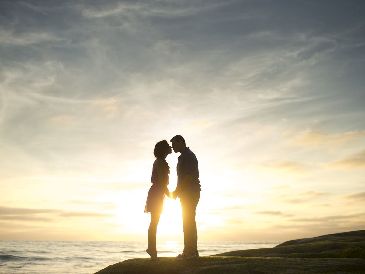 A couple is silhouetted against the setting sun, standing close together on a beach with a serene ocean and a dramatic sky in the background.