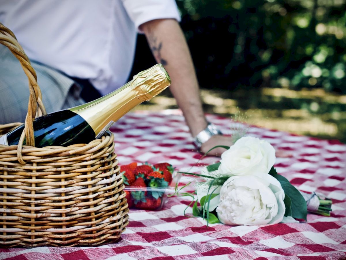 A picnic setup with a basket holding a bottle of champagne, a bouquet of white roses, and a container of strawberries on a checkered tablecloth.