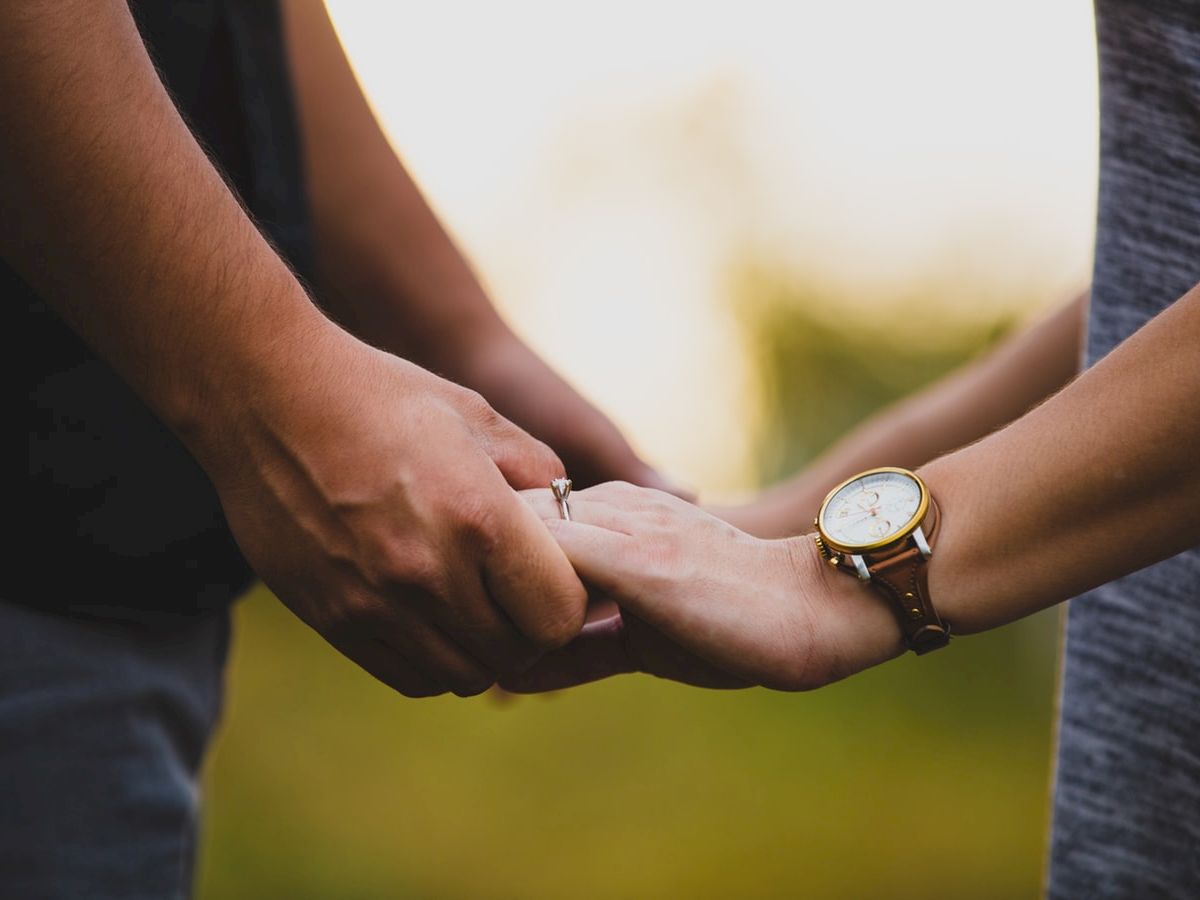 A close-up image shows two people holding hands outdoors, one person is wearing a wristwatch and an engagement ring on their finger.