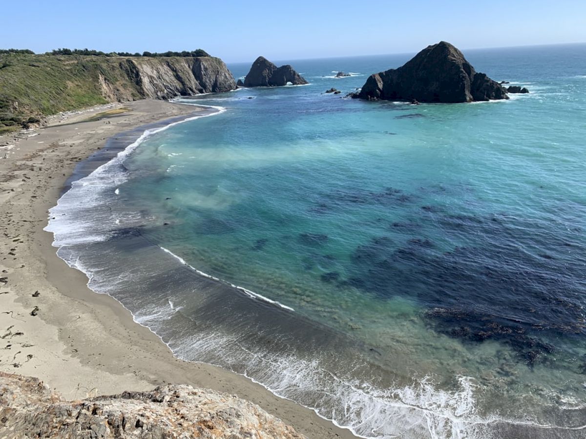 A scenic coastal view with a sandy beach, clear blue water, rocky cliffs, and a few large rocks or small islands near the shore, under a clear sky.