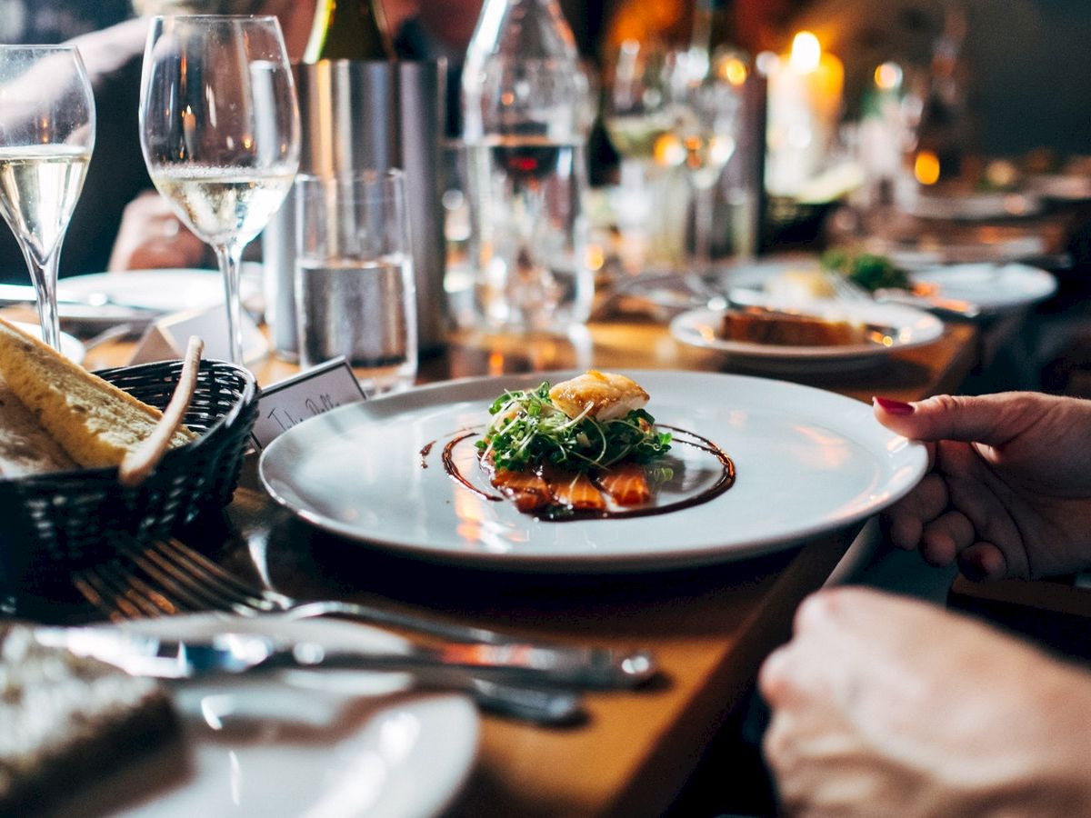 The image shows a fine dining setup with a plated dish, bread basket, glasses of wine, and cutlery, depicting a sophisticated dinner setting.